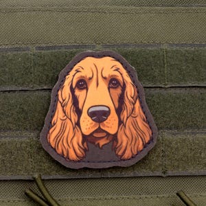 Puede incluir: Un parche con una ilustración detallada de la cara de un perro Cocker Spaniel, con pelaje marrón y naranja, una nariz negra y ojos expresivos. El parche está adherido a un fondo de tela verde.