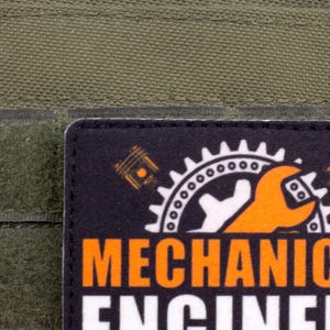 Pu&ograve; includere: Una toppa nera con le parole "MECHANICAL ENGINEER" in bianco e arancione. La toppa presenta un ingranaggio, una chiave inglese e un pistone. Lo sfondo &egrave; verde oliva.
