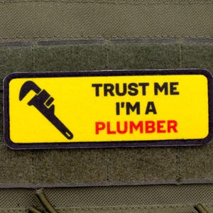 May include: A rectangular patch with a yellow background and black border. The patch features a black wrench graphic and the text "TRUST ME I'M A PLUMBER" in black and red. The patch is set against a green background.