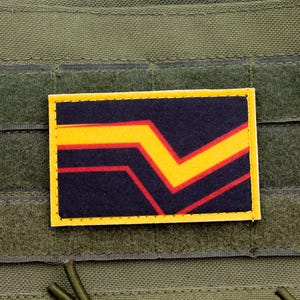 Op de afbeelding: Een rechthoekige patch met een gele rand en een ontwerp met een gele chevron en rode strepen op een marineblauwe achtergrond. De patch is bevestigd op een groene stoffen ondergrond.