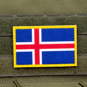 May include: A rectangular patch featuring the flag of Iceland, with a blue background, a red cross, and white borders. The patch is framed with a yellow border and is affixed to a green fabric surface.