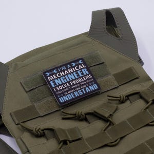 May include: Olive green tactical vest with a patch that reads "I'M A MECHANICAL ENGINEER I SOLVE PROBLEMS YOU DON'T KNOW YOU HAVE IN WAYS YOU CAN'T UNDERSTAND". The vest has multiple straps and attachment points.