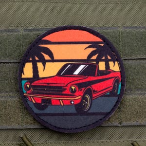 Puede incluir: Parche redondo con un coche descapotable rojo con detalles negros, sobre un fondo de puesta de sol con palmeras. El diseño es circular y tiene un borde negro. El parche está fijado a una tela verde.