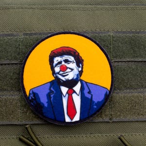 May include: A round yellow patch with a blue and white image of a man with a red clown nose. The man is wearing a blue suit and a red tie. The patch is attached to a green fabric background.
