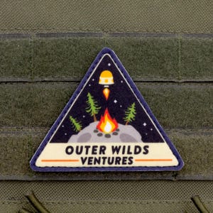 Może przedstawiać: Tr&oacute;jkątna naszywka o tematyce kosmicznej i kempingowej. Naszywka przedstawia rakietę, ognisko, drzewa i napis "OUTER WILDS VENTURES" białymi literami. Naszywka jest umieszczona na ciemnoniebieskim tle.