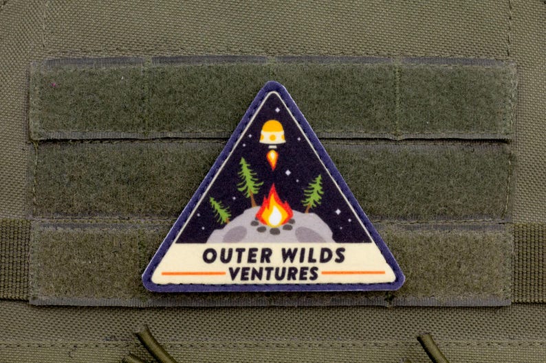 Może przedstawiać: Tr&oacute;jkątna naszywka o tematyce kosmicznej i kempingowej. Naszywka przedstawia rakietę, ognisko, drzewa i napis "OUTER WILDS VENTURES" białymi literami. Naszywka jest umieszczona na ciemnoniebieskim tle.