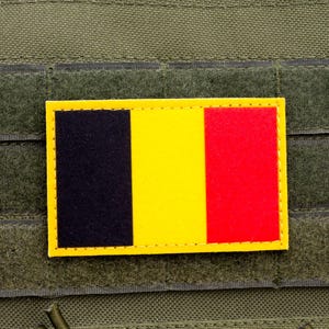 以下が含まれることがあります： ベルギーの国旗をあしらった長方形のパッチ。黒、黄、赤の縦縞模様です。パッチは黄色の縁取りがあり、緑色の布の背景に固定されています。