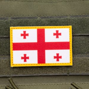 May include: A white flag with a red cross and four smaller red crosses on a green background. The flag is sewn onto a piece of fabric.