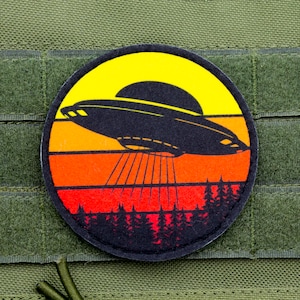 May include: A round black patch with a retro sunset design featuring a flying saucer in silhouette. The patch has a hook and loop backing for attaching to clothing or bags.