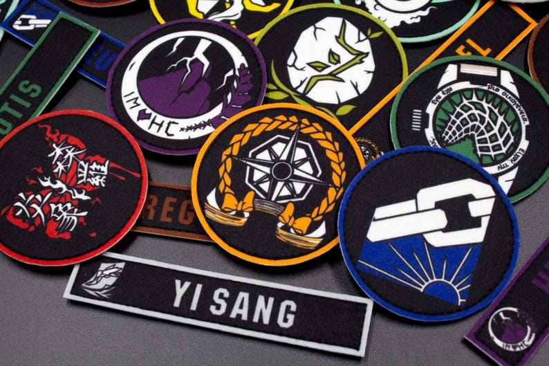 Puede incluir: Una colecci&oacute;n de parches bordados con varios dise&ntilde;os, incluyendo una br&uacute;jula, un eslab&oacute;n de cadena, una planta y una criatura con una telara&ntilde;a. Los parches son circulares y tienen colores negro, blanco, azul, verde y naranja. El texto "Yi Sang" es visible en un parche negro.