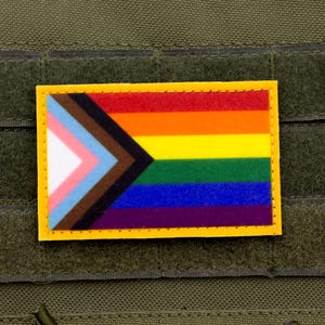 May include: A rectangular patch featuring the Progress Pride flag with a yellow border. The flag includes the traditional rainbow stripes, along with a chevron of colors representing transgender, Black, and brown communities. The patch is set against a green background.