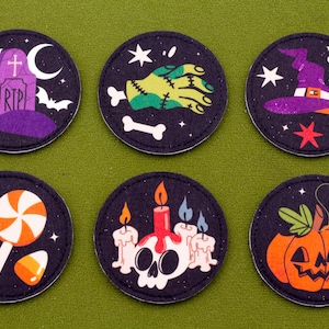 Op de afbeelding: Set van zes ronde Halloween-patches. Elke patch heeft een ander ontwerp: een grafsteen, een zombiehand, een heksenhoed, snoepmaïs, een schedel met kaarsen en een pompoen. De patches zijn zwart met witte stiksels.