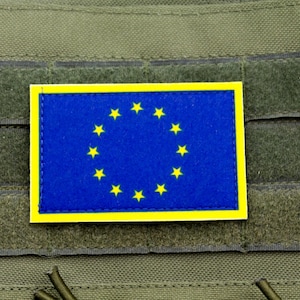 以下が含まれることがあります： 欧州連合の旗が描かれた長方形のパッチ。旗は青色の背景に12個の黄色の星が円を描いています。パッチは黄色の縁取りがあり、緑色の生地に固定されています。