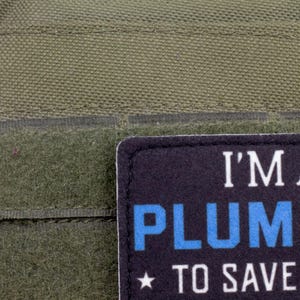 K&ouml;nnte beinhalten: Nahaufnahme eines dunkelgr&uuml;nen Stoffes mit einem rechteckigen Aufn&auml;her. Der Aufn&auml;her ist schwarz mit wei&szlig;em und blauem Text, der lautet: "I'M A PLUMBER TO SAVE TIME". Unter dem Text befindet sich ein kleiner Stern.