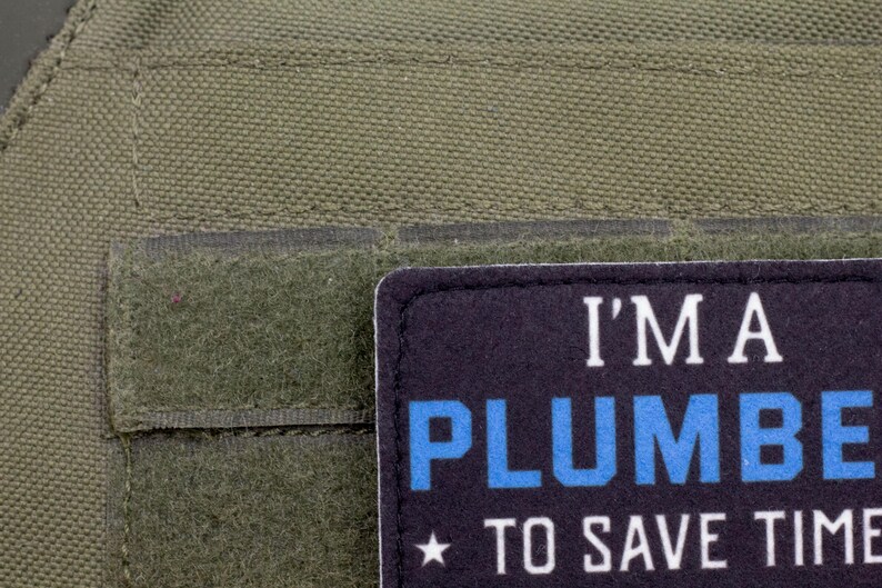 K&ouml;nnte beinhalten: Nahaufnahme eines dunkelgr&uuml;nen Stoffes mit einem rechteckigen Aufn&auml;her. Der Aufn&auml;her ist schwarz mit wei&szlig;em und blauem Text, der lautet: "I'M A PLUMBER TO SAVE TIME". Unter dem Text befindet sich ein kleiner Stern.