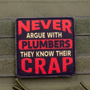 Op de afbeelding: Een rechthoekige patch met de tekst "NEVER ARGUE WITH PLUMBERS THEY KNOW THEIR CRAP" in rode en witte letters op een zwarte achtergrond. De patch is bevestigd aan een groene stof.