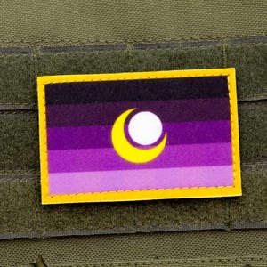 Op de afbeelding: Een rechthoekige patch met een gele rand, met een ontwerp van een halve maan en een witte cirkel op een achtergrond van horizontale paarse strepen. De patch is bevestigd op een groene stof.