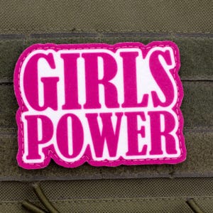 May include: A rectangular patch with the words "GIRLS POWER" in bright pink, outlined in white. The patch is attached to a green fabric background with a hook and loop fastener.