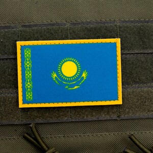 May include: A rectangular patch displaying the flag of Kazakhstan, with a blue field, a yellow sun, and a golden eagle. The patch has a yellow border and is attached to a green tactical vest.