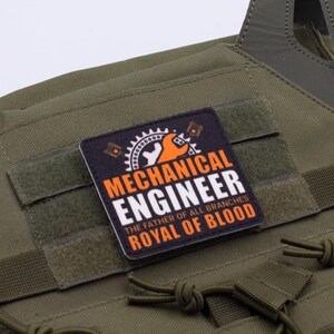 Pu&ograve; includere: Gilet tattico verde oliva con una toppa rettangolare. La toppa presenta le parole "MECHANICAL ENGINEER" in bianco e arancione, insieme al testo "THE FATHER OF ALL BRANCHES ROYAL OF BLOOD" e una grafica di ingranaggi e chiave.