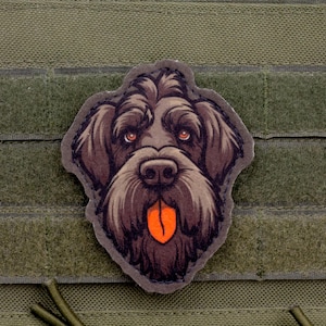 May include: A patch featuring a detailed illustration of a dog's face with brown fur, orange eyes, and a protruding orange tongue. The patch is set against a green background with a hook and loop fastener.