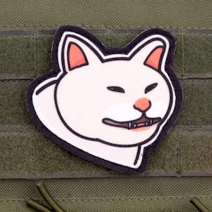 Può includere: Una toppa a forma di gatto bianco con orecchie interne e naso rosa, caratterizzata da un'espressione facciale compiaciuta con occhi socchiusi e una piccola bocca che mostra due denti. La toppa ha un bordo nero spesso su un tessuto verde oliva.