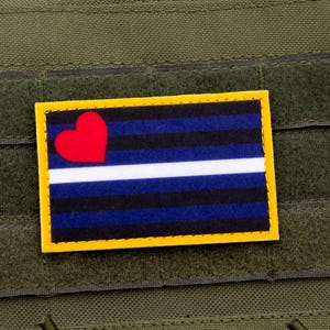 Op de afbeelding: Een zwarte, blauwe en witte patch met een rood hart in de linkerbovenhoek. De patch heeft een gele rand en is bevestigd aan een groene stoffen achtergrond.
