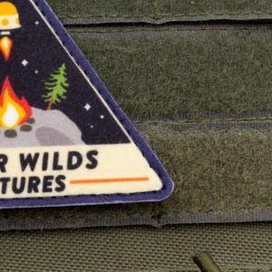 Może przedstawiać: Tr&oacute;jkątna naszywka o tematyce kosmicznej, przedstawiająca ognisko, drzewa i rakietę. Naszywka ma napis "OUTER WILDS VENTURES" białymi literami z pomarańczowym podkreśleniem, na ciemnoniebieskim tle. Naszywka jest na zielonej tkaninie.