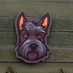 Op de afbeelding: Een rechthoekig olijfgroen tactisch vestpaneel met een centraal geplaatste, uitgestanste morale patch. De patch toont de kop van een donkerharige Schotse Terriër met puntige, oranje omrande oren en amberkleurige ogen in een grafische illustratiestijl.