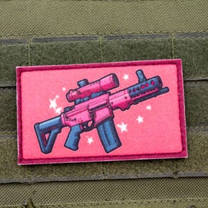 Può includere: Un fucile rosa e nero con un cannocchiale e stelle su uno sfondo rosa. Il fucile è su una toppa.