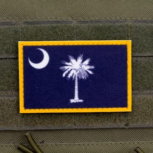 May include: A rectangular patch featuring the South Carolina state flag. The flag has a dark blue background with a white crescent moon in the upper left corner and a white palmetto tree in the center. The patch is bordered with a yellow frame.