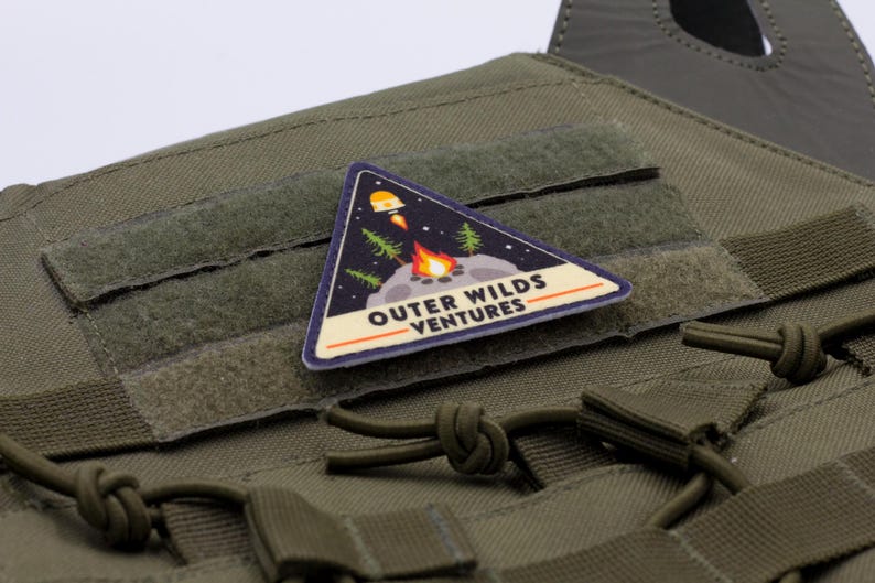 Może przedstawiać: Tr&oacute;jkątna naszywka z napisem "OUTER WILDS VENTURES" oraz grafiką rakiety, ogniska i drzew. Naszywka znajduje się na zielonej tkaninie z rzepem i paskami. Naszywka ma szerokość około 5 cm.