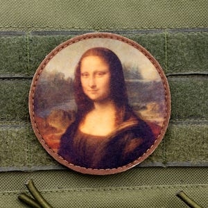 Pode incluir: Um remendo redondo de cor castanho com uma borda costurada apresenta uma imagem impressa da Mona Lisa. A imagem está colocada sobre um fundo pintado de uma paisagem com um rio e árvores.