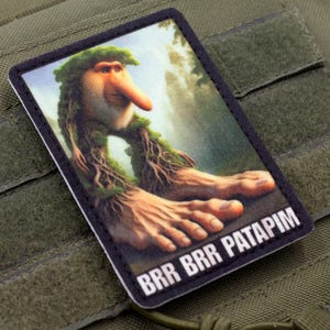 May include: A rectangular patch featuring a whimsical illustration of a creature with a long nose, leafy hair, and large feet. The text "BRR BRR PATAPIM" is printed at the bottom. The patch is set against a green fabric background.