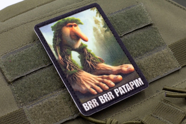 May include: A rectangular patch featuring a whimsical illustration of a creature with a long nose, leafy hair, and large feet. The text "BRR BRR PATAPIM" is printed at the bottom. The patch is set against a green fabric background.