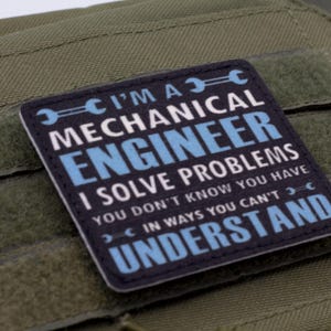 May include: A square patch with the text "I'M A MECHANICAL ENGINEER I SOLVE PROBLEMS YOU DON'T KNOW YOU HAVE IN WAYS YOU CAN'T UNDERSTAND" in white and blue on a black background. The patch is on a green fabric.