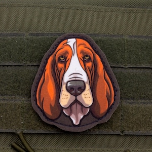 Könnte beinhalten: Ein Aufnäher mit einer detaillierten Illustration eines Basset Hound-Kopfes. Der Hund hat braunes und weißes Fell, lange, hängende Ohren und ein schlaffes Gesicht. Der Aufnäher ist auf einem grünen Stoffhintergrund angebracht.