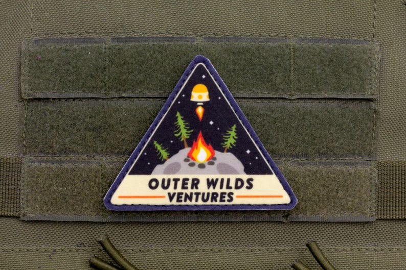 Może przedstawiać: Tr&oacute;jkątna naszywka o tematyce kosmicznej i kempingowej. Naszywka przedstawia rakietę startującą z planety, ognisko i sosny na tle gwiaździstego nieba. Tekst "OUTER WILDS VENTURES" jest wyświetlany na biało.