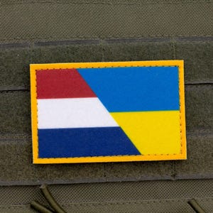 May include: A rectangular patch with a combined flag design. The patch features the colours of the Dutch and Ukrainian flags. The Dutch flag is red, white, and blue, while the Ukrainian flag is blue and yellow. The patch has a yellow border.