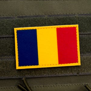 May include: A rectangular patch featuring the Romanian flag, with vertical stripes of blue, yellow, and red, set against a yellow border. The patch is affixed to a dark green, textured fabric.