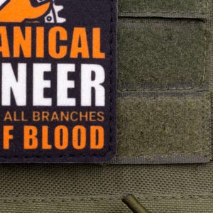 Pu&ograve; includere: Primo piano di una toppa con le parole "MECHANICAL ENGINEER" in lettere arancioni e bianche. La toppa &egrave; su uno sfondo di tessuto verde. &Egrave; visibile anche il testo "FATHER OF ALL BRANCHES" e "SEAL OF BLOOD".
