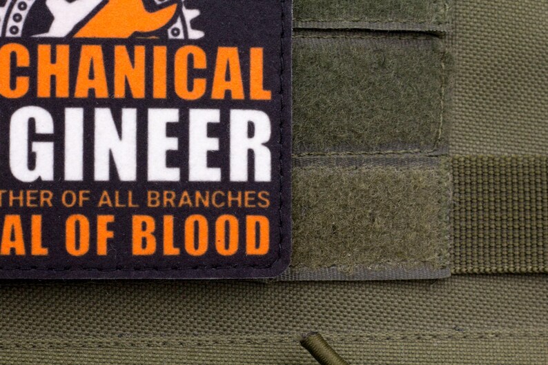 Pu&ograve; includere: Primo piano di una toppa con le parole "MECHANICAL ENGINEER" in lettere arancioni e bianche. La toppa &egrave; su uno sfondo di tessuto verde. &Egrave; visibile anche il testo "FATHER OF ALL BRANCHES" e "SEAL OF BLOOD".