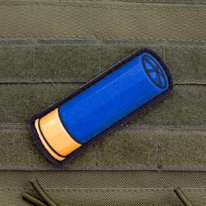 May include: A blue and yellow shotgun shell patch with a black border is affixed to a green tactical vest. The patch features a blue body and a yellow base, with a black design on the top.