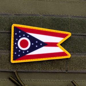 May include: A patch featuring the flag of Ohio, with a yellow border. The flag design includes a blue triangle with white stars, a red circle, and red and white stripes. The patch is attached to a green fabric.