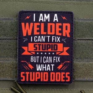 Op de afbeelding: Een rechthoekige zwarte patch met afgeronde hoeken voorzien van rode en witte tekst die luidt I AM A WELDER I CAN'T FIX STUPID BUT I CAN FIX WHAT STUPID DOES, geaccentueerd met kleine bliksem- en lastoortsiconen, bevestigd op olijfgroene stof.