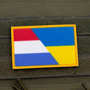 May include: A rectangular patch with a yellow border, displaying the flags of the Netherlands and Ukraine. The Netherlands flag has red, white, and blue stripes. The Ukrainian flag is blue and yellow. The patch is on green fabric.
