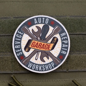 May include: A round patch with the words "AUTO REPAIR GARAGE WORKSHOP SERVICE" encircling crossed wrenches and a screwdriver. The patch is white with a dark blue border and red star accents. It is affixed to a green fabric.