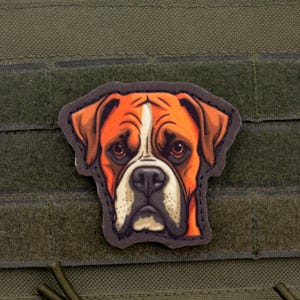Pode incluir: Um patch com o rosto de um cão Boxer, com um esquema de cores castanho e branco. O patch é fixado a um fundo de tecido verde. O rosto do cão é detalhado com uma faixa branca no nariz e uma expressão séria.