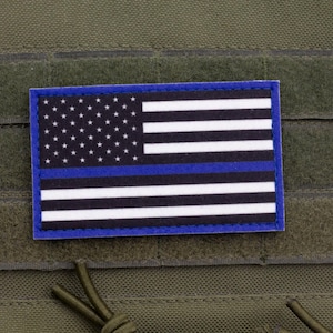 Puede incluir: Un parche rectangular con la bandera estadounidense, con un borde azul y una línea azul. La bandera tiene rayas blancas y un campo de estrellas. El parche está fijado a un fondo de tela verde.