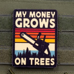 Könnte beinhalten: Rechteckiger Aufnäher mit dem Text "MY MONEY GROWS ON TREES" in weißen Buchstaben. Das Design umfasst eine Kettensägen-Silhouette, einen farbenfrohen Sonnenuntergang und eine Waldszene. Der Aufnäher ist an einem grünen Stoff befestigt.